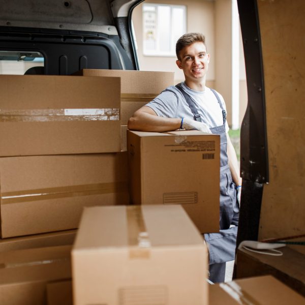 A young handsome smiling worker wearing uniform is standing next to the van full of boxes. House move, mover service.