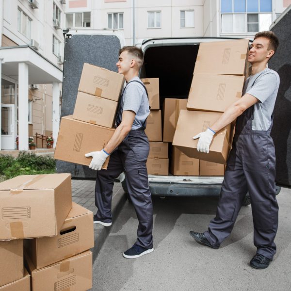 Two young handsome smiling workers wearing uniforms are unloading the van full of boxes. The block of flats is in the background. House move, mover service.