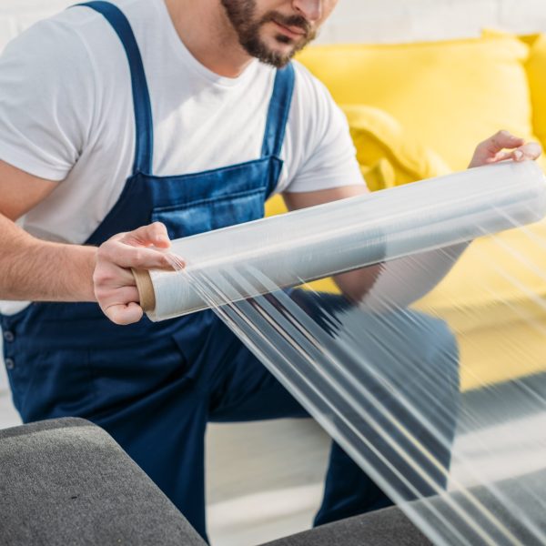 cropped view of mover wrapping furniture with roll of stretch film in apartment